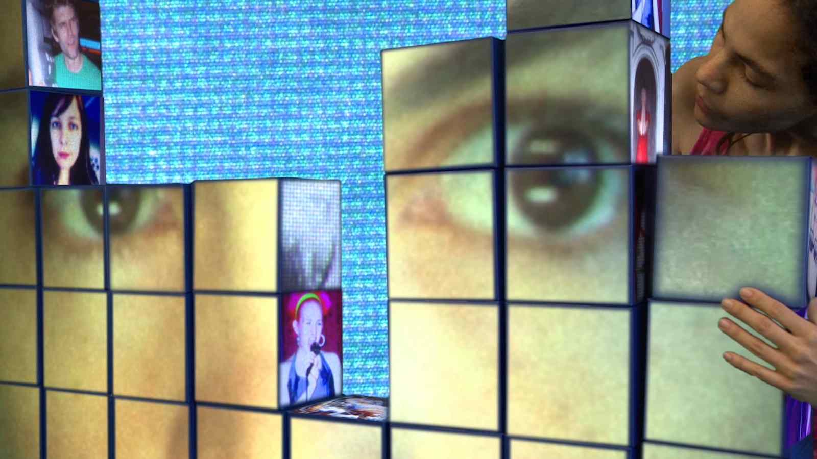 A woman is standing in front of a wall of cubes with faces on them.