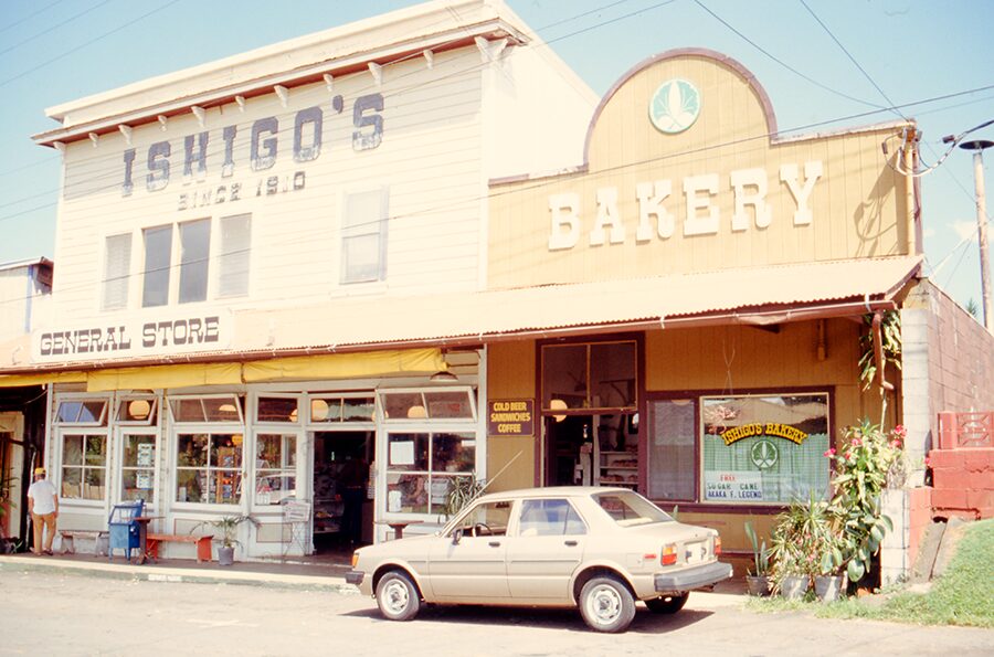 tan car parked on street in front of storefronts including a bakery. 1950's in California.