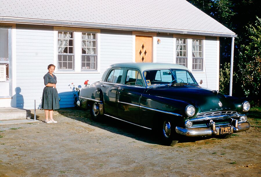 woman wearing blue dress and white shoes outside ranch home standing next to a dodge car 1950s.